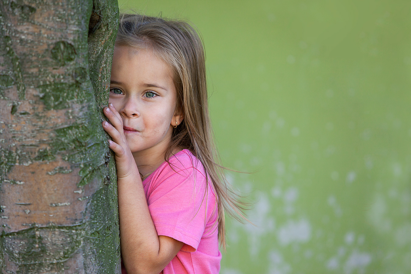 Fotografie eines Mädchens im rosa T-Shirt, hald hinter einem Baum versteckt und hervorlugend